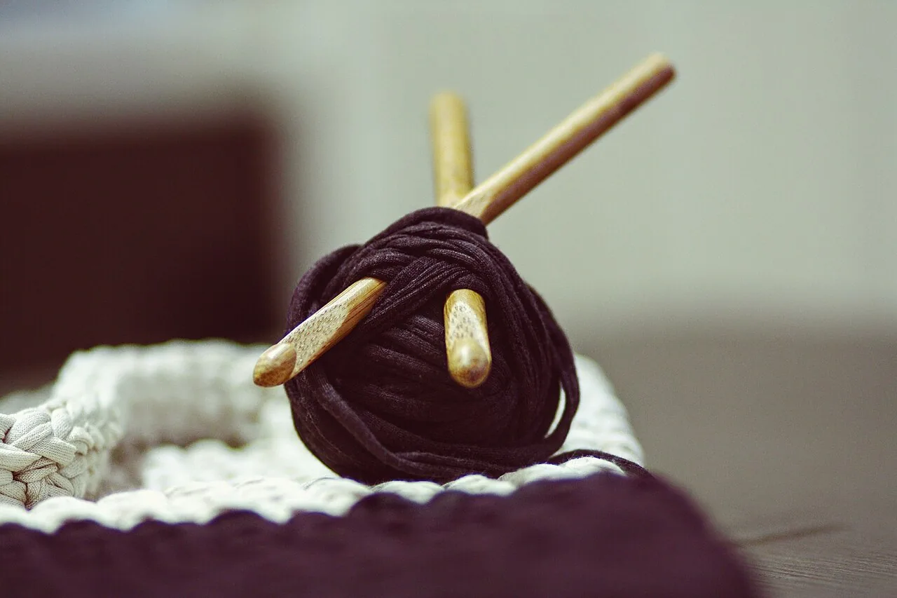 Wooden knitting needles crossing through a ball of deep purple yarn beside knitted fabric.